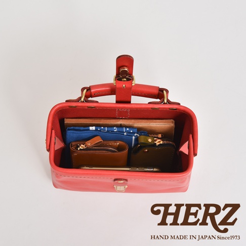 職人手作紅色皮革迷你軟皮杜勒斯包，包內裝有藍色印花布、棕色與深綠色皮夾，以及一支手機。包包採用金屬釦件與提把設計。