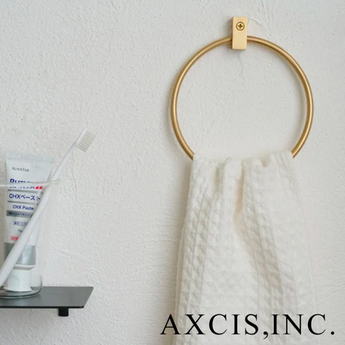 黃銅圓形毛巾環，表面光滑，呈金色。毛巾環固定在牆壁上，並掛有一條白色格紋毛巾。左側有一個黑色架子，上面放著牙膏管和牙刷。