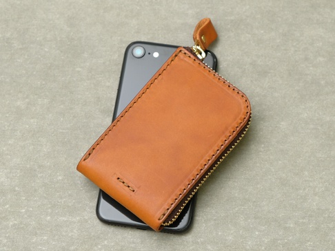 職人手作皮革Organ全包式拉鍊卡片鑰匙包，棕色皮革，拉鍊開合，附有皮革拉環，放置於黑色手機上方。