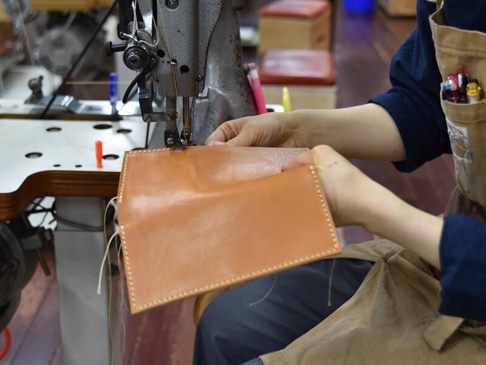 職人正在使用縫紉機縫製一個淺棕色皮革扁長夾，展現其簡約的皮革紋理與精緻的車縫細節。