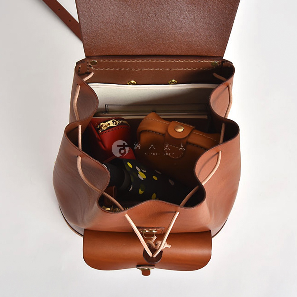 職人手作的棕色皮革後背包，包內裝有紅色零錢包、棕色皮夾及花紋摺疊傘，包口以束繩和金屬扣固定，展現經典簡約的設計。