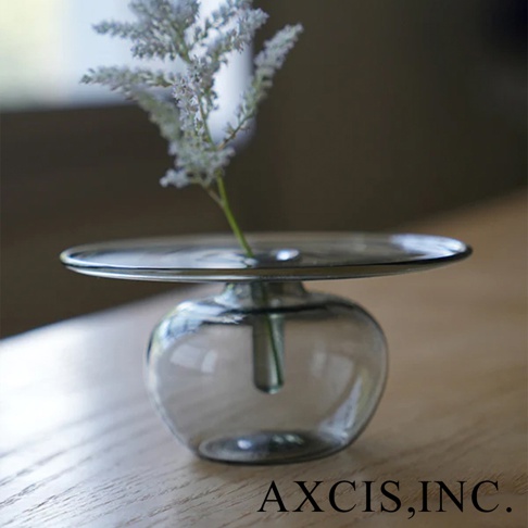 AXCIS, INC. 的 Lua 線香玻璃花器，底部為圓潤的透明玻璃，上方為扁平的灰色玻璃盤，盤中插有一束白色細碎的小花。