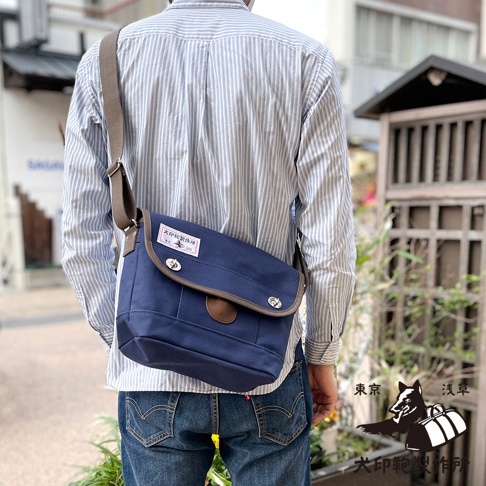 深藍色純棉帆布郵差包，有棕色飾邊和翻蓋，配有兩個金屬扣和一個皮質圓形標籤。包帶為棕色。