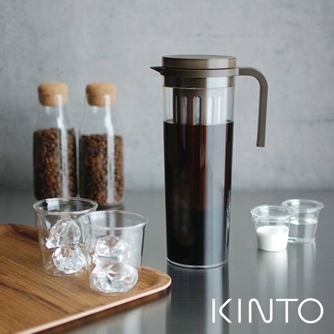 PLUG 咖啡冷泡壺，透明玻璃壺身，裝有深棕色冷泡咖啡，搭配深灰色壺蓋與提把。右側有兩個透明小杯，分別裝有牛奶與水。左側為兩個裝有冰塊的玻璃杯，以及兩瓶裝有咖啡豆的玻璃瓶。