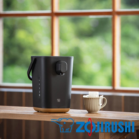 STAN美型微電腦熱水瓶，黑色霧面機身搭配大地色系底座，設有提把和出水口，側面有水位顯示刻度。旁邊擺放著一個淺米色帶有紋路的杯子，杯內放有茶包。