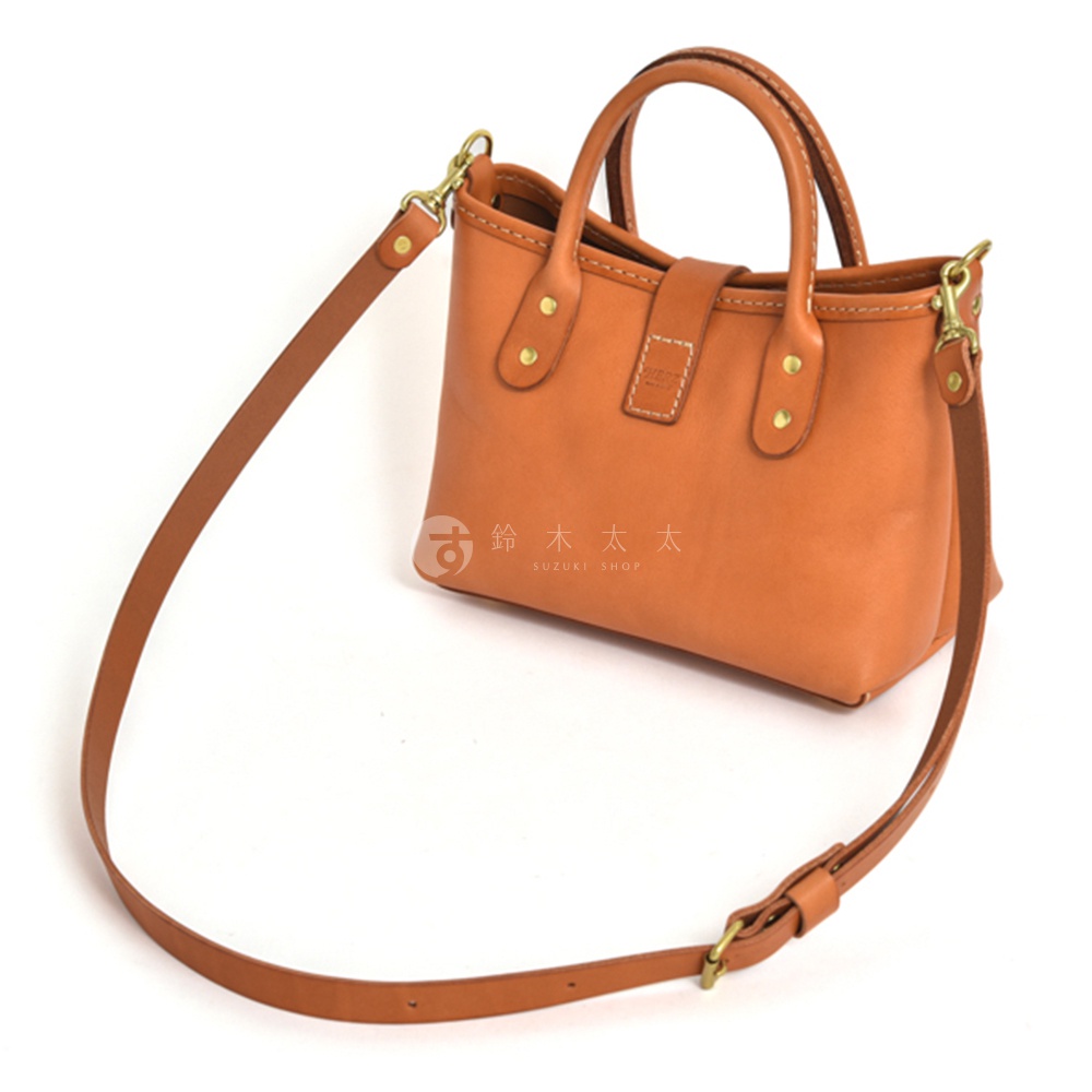 職人手作皮革迷你托特包，呈現經典的棕褐色調。包身使用柔軟的皮革製成，配有雙提把和可拆卸肩帶，方便肩背或手提。包身結構簡潔，綴有金屬鉚釘和皮革搭扣，展現精緻工藝。