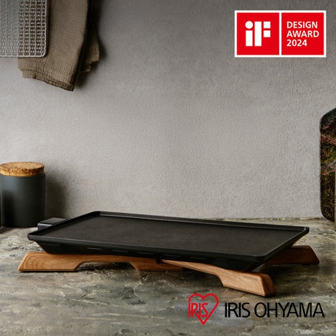 黑色輕薄電烤盤，表面為黑色鑄鐵材質，設有兩個木製把手。烤盤放置在兩個木製支架上，背景為灰色紋理牆面。