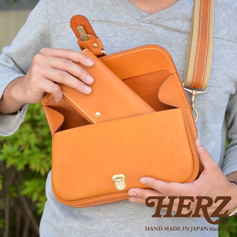 一隻由職人手作的橘棕色皮革單扣斜背包，包身有縫線裝飾，可見包內有一個長夾，包的正面有一個金色單扣。