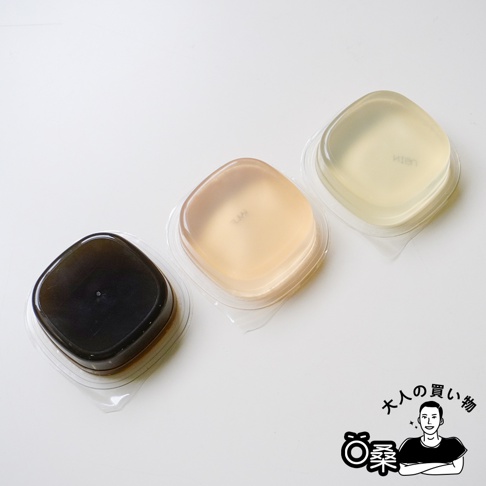 三個獨立包裝的半透明方形甜點排成一列，由左至右分別為深色、淺琥珀色與透明色，放置於白色背景上，右下角有一位卡通人物的黑白插畫與文字裝飾。
