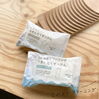 兩塊白色包裝的洗衣皂，上面印有日文說明和衣物圖示，置於淺色木紋背景上，背景後方可見一個木製的洗滌板。