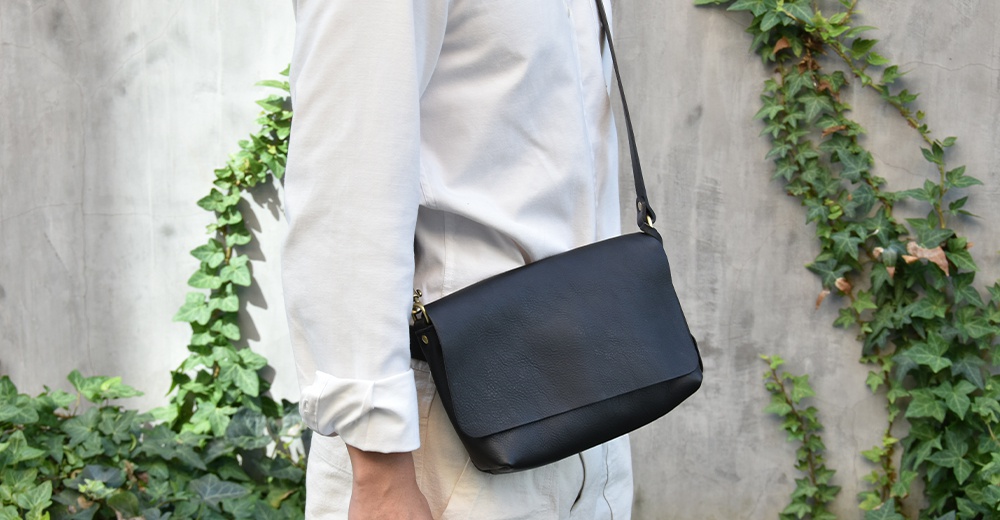 一個人斜背著一個黑色皮革隨身包，包包有翻蓋設計，背景是佈滿爬藤植物的灰色水泥牆。