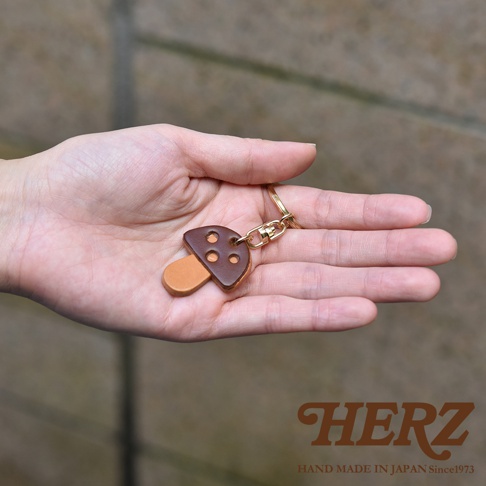 職人手作皮革香菇造型鑰匙圈，蘑菇傘為咖啡色皮革，傘柄為淺棕色皮革，傘面有三個圓形裝飾孔，搭配金色鑰匙圈。