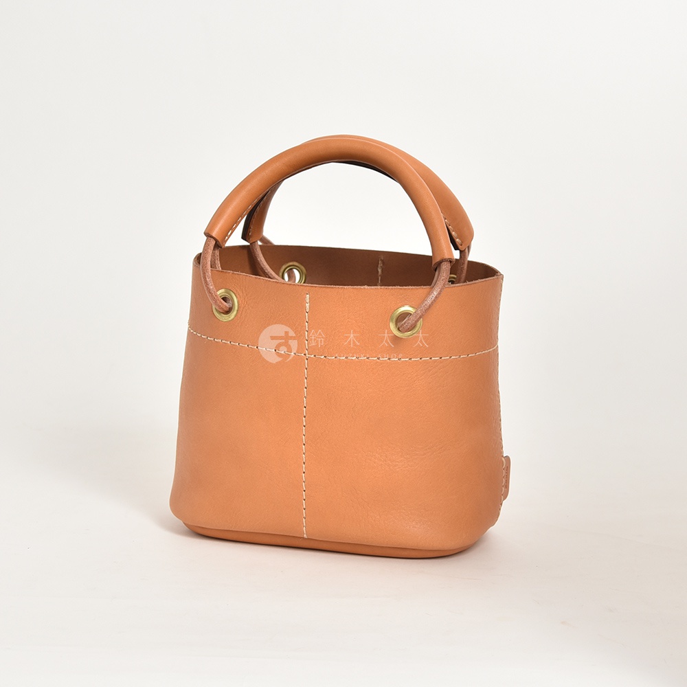 一個棕褐色皮革製成的手提午餐袋，有著加厚的圓形提把，袋口處有金屬孔和抽繩設計，縫線明顯，風格簡潔。
