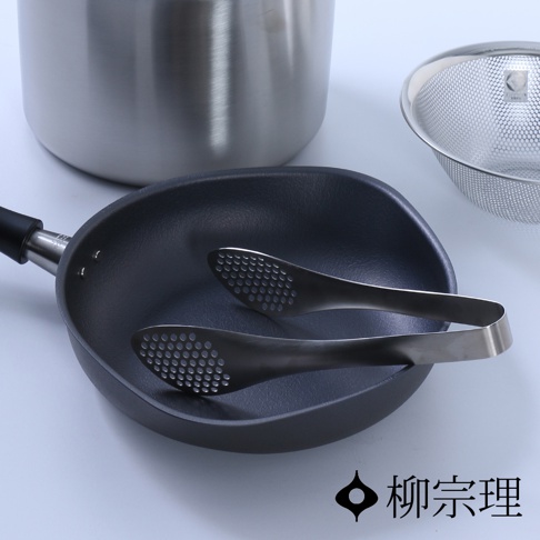 柳宗理廚具組合包含一個帶有金屬蓋的亮面不鏽鋼鍋、一個不鏽鋼細網漏勺，以及一個放置在深灰色不沾平底鍋內的打孔不鏽鋼料理夾，整體呈現簡約的工業設計風格。