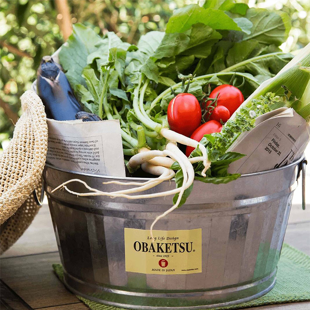 一個裝滿新鮮蔬菜的金屬桶，桶上貼有「OBAKETSU」品牌標籤，標籤上寫著「MADE IN JAPAN」。