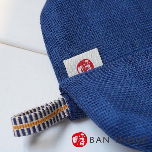 藍色蚊帳質地防燙鍋套，帶有白色織標，標籤上有紅色圖案。鍋套一側有一個藍白黃條紋編織提帶。