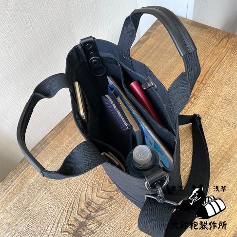 黑色帆布托特包，內部裝有手機、錢包、筆記本、紅色筆和一瓶水。包有兩個提把和一條可拆卸的斜背包帶。