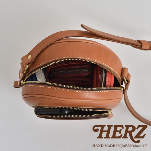 職人手作皮革小圓包，採用鞣製皮革製成，顏色為溫潤的棕色。包身呈現圓潤飽滿的造型，並配有拉鍊開闔。包內可見紅色錢包及手機等物品。