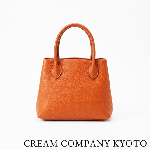 這是一款由京都品牌 CREAM COMPANY KYOTO 推出的橘色手提托特包，採用輕量耐用的 glove 牛皮革製作，外觀呈現細緻的皮革紋理，包身簡約精巧，手柄設計穩固，下方標註有品牌名稱，適合日常休閒攜帶。