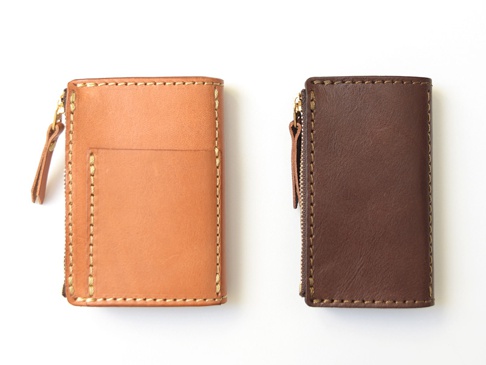 兩件職人手作皮革拉鍊零錢鑰匙包，左為淺棕色，右為深棕色。兩款皆為直立式設計，使用金屬拉鍊，並以手工縫線裝飾。淺棕色零錢包外觀有一個方形插槽，深棕色零錢包則為素面設計。