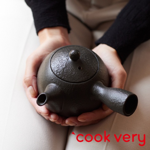 雙手捧著一個深灰色、帶有紋理的常滑燒咖啡急須茶壺，壺身飽滿，設有彎曲的壺嘴和手柄，壺蓋中央有圓形握鈕。