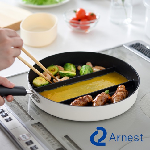 這是一個放置在電磁爐上的Arnest三合一分隔不沾平底鍋，鍋面被隔板劃分為三個區域，畫面中一隻手正使用木製筷子翻動左側的蔬菜與蝦仁，中央長形區域正在烹飪蛋液，右側則是煎煮好的肉捲，整體呈現出高效便捷的早餐或便當烹飪場景。