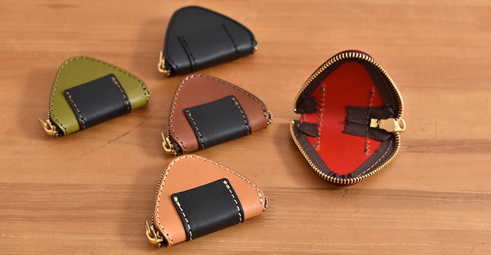 桌面上擺放著五個日本HERZ職人手作的三角飯糰造型零錢包，包含黑色、橄欖綠、棕色、淺棕色及一個內部呈現紅色內襯的開口展示款，每個零錢包皆由質感皮革製成並飾有對比色的黑色皮件細節與金色拉鍊。