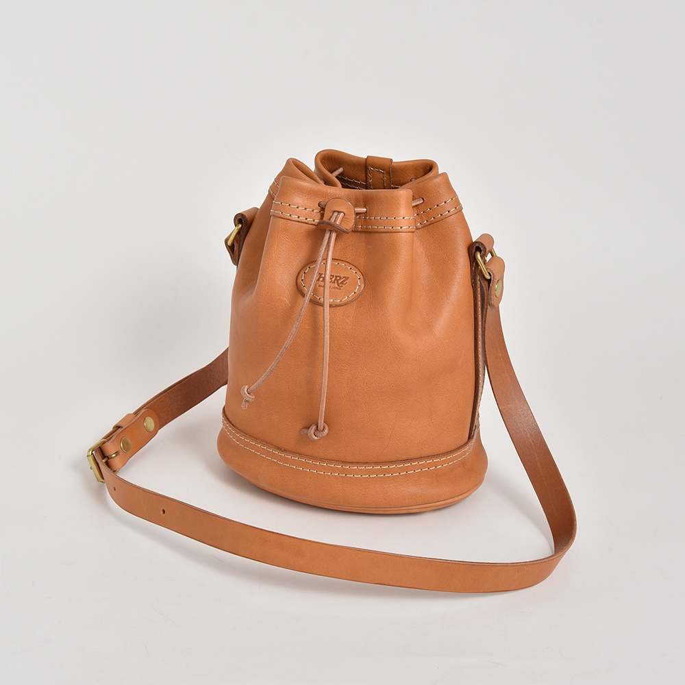 淺棕色皮革束口單肩包，圓筒形設計，附有可調節的皮革背帶和束繩。包身中央有一個橢圓形皮革標誌，印有品牌名稱。