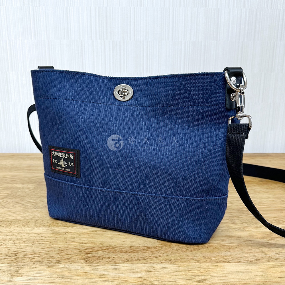 深藍色刺子織迷你側背包，帶有菱形紋理，銀色旋轉鎖扣，黑色織帶肩帶，側邊有紅色標籤。