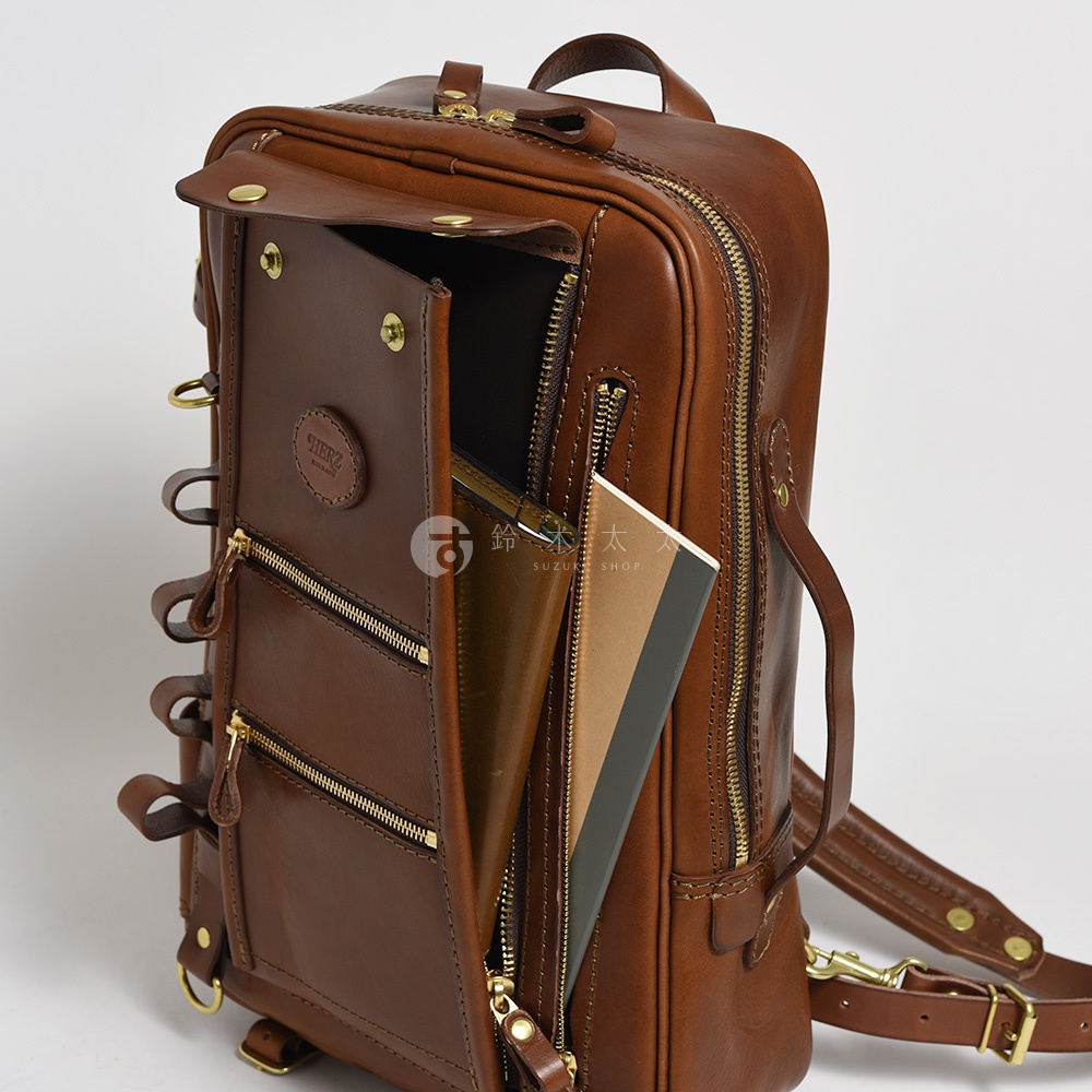 職人手作棕色皮革兩用多功能後背包，掀蓋設計，前方有三個拉鍊口袋，側邊有金屬 D 型環，內部可見筆記本和錢包。