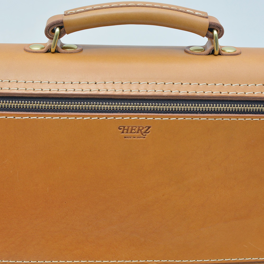 職人手作皮革旅行商務兩用波士頓包，展示包款正面上半部，淺棕色皮革，配有黃銅色金屬扣環和黑色拉鍊，頂部有皮革提把，中央印有「HERZ」品牌標誌。