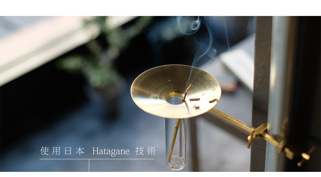 黃銅製的線香架，頂部有一個圓形碟狀的集灰盤，盤上插有燃燒中的線香，並冒出裊裊白煙。線香架透過夾子固定在深色支架上，底部連接一個透明玻璃試管，試管內有少量灰燼。