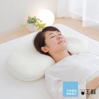 一位女性仰臥在白色床鋪上，頭部枕著一個白色、柔軟的雲狀枕頭。枕頭呈彎月形，表面光滑，材質看似布料。背景有植物與圓形燈飾。