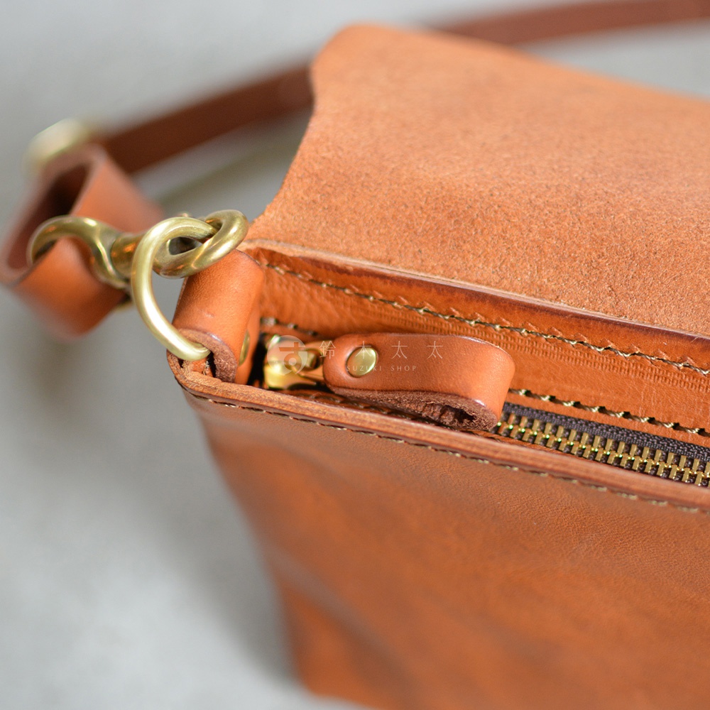 特寫鏡頭展示了棕褐色皮革斜背包的細節，包括黃銅色的拉鍊、皮製拉鍊頭以及連接肩帶的金屬環扣。
