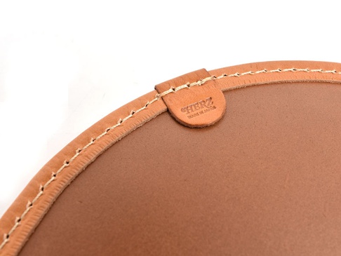 職人手作皮革圓形坐墊，採用棕色皮革材質，邊緣以米白色縫線手工縫製，並有一個帶有「HERZ MADE IN JAPAN」字樣的皮革吊牌。