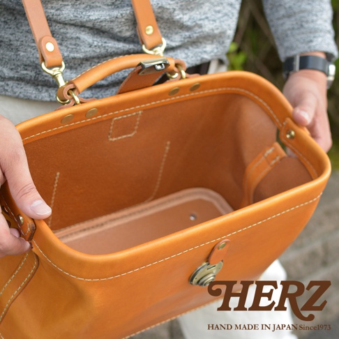 職人手作的皮革長型迷你醫生包，呈現溫暖的淺棕色調，材質為真皮。包包結構紮實，縫線清晰，配有金屬鎖扣和皮質提把，可調節的肩帶也由皮革製成，展現精緻的手工藝。