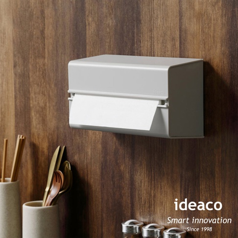 灰白色ABS材質的壁掛/桌上兩用擦手紙架，設計簡潔，可容納紙巾，掛在木紋牆面上，下方擺放餐具。