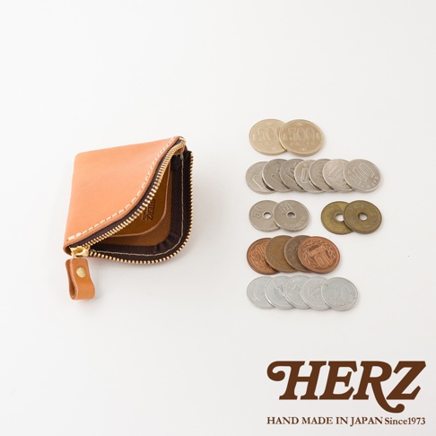 一張從上方拍攝的平鋪照片，左側為一個小巧的淺棕色皮革零錢包，採L型拉鍊設計，配有帶金屬扣的皮革拉鍊頭，內部有隔層；右側整齊排列著多枚日本硬幣，下方印有HERZ HAND MADE IN JAPAN Since1973的咖啡色品牌標誌。
