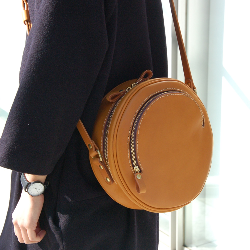 職人手作皮革小圓包，採用天然皮革製作，呈現溫潤的焦糖棕色。包身呈圓形，雙層拉鍊設計，搭配細緻縫線與皮革拉頭，風格簡約復古。