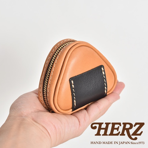 職人手作皮革三角飯糰造型小物收納包，以淺棕色皮革製成，搭配黑色皮革飾片，並以白色縫線點綴。包包設計成可愛的三角飯糰形狀，側邊有金色拉鍊，整體風格簡約實用。