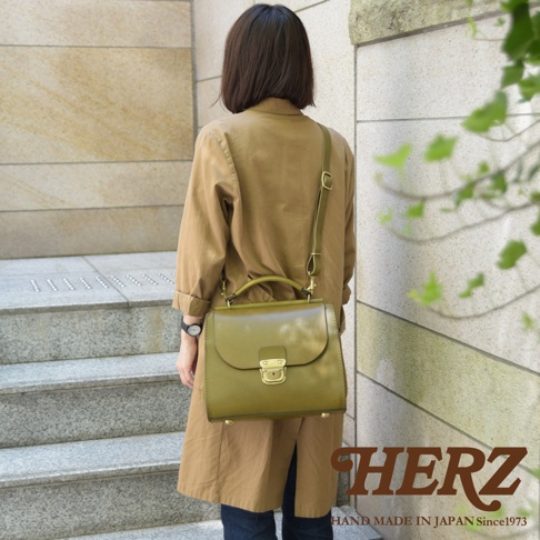 日本HERZ職人手作皮革屋瓦風兩用單肩包，女性背對鏡頭，身穿卡其色風衣，斜背著橄欖綠色皮革包，包上有金色鎖扣，下方壓印HERZ HAND MADE IN JAPAN Since1973字樣。