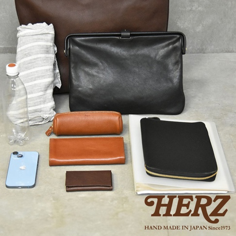 圖片中展示了多款皮革製品，包括一個黑色手拿包、一個棕色長夾、一個棕色筆袋、一個深棕色卡夾，以及一個淺藍色手機。背景還有一個棕色托特包，左側放著一瓶水和一條條紋毛巾。圖片右下角有HERZ的品牌字樣。