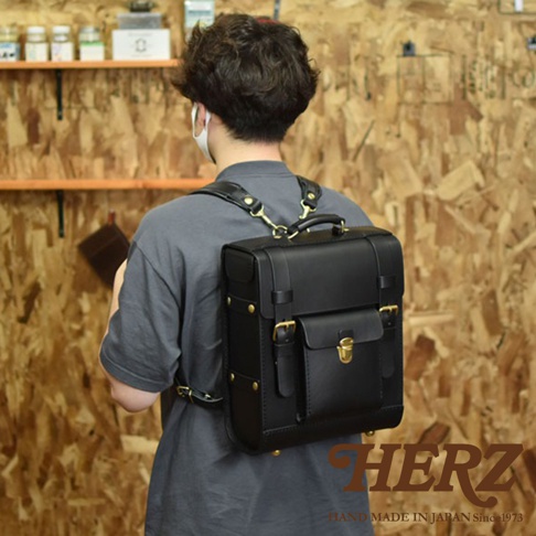 一位男士背著一個黑色皮革經典學院風後背包 R-100。背包為方形設計，採用全皮革材質，配有黃銅色金屬扣環、鎖扣及鉚釘裝飾。背包的正面有一個翻蓋小口袋，上方有提把，肩帶為寬版皮質設計。