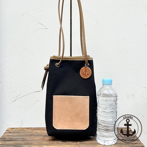 神戶clueto黑色束口肩背包，配有大地色皮革口袋，附有錨形吊飾，旁邊放著一瓶水。