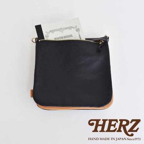 黑色皮革製作的直式斜背包，底部有天然皮革的翻邊設計，搭配金色拉鍊及吊環，內側露出筆記本一角。