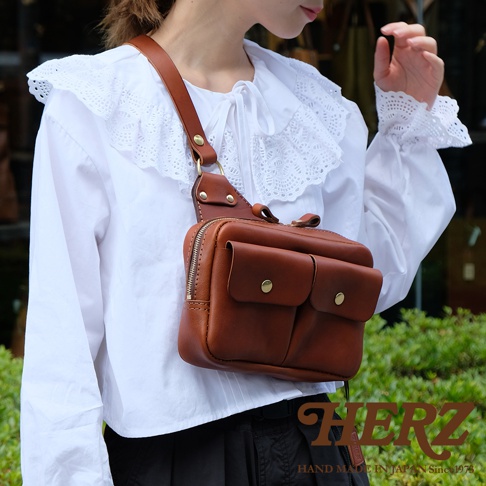 棕色皮革雙口袋斜背包，搭配金色金屬釦，配戴於身穿白色蕾絲領襯衫的模特兒身上。