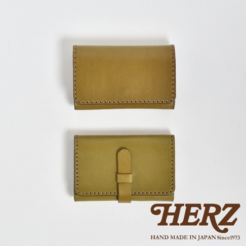 這張照片呈現日本 HERZ 職人手作的經典皮革名片夾，共有兩款展示。上方為簡潔的無扣設計，下方則附有皮革插扣固定，兩者皆採用溫潤的橄欖綠色手工縫製而成，展現細膩的皮革質感與專業工藝，右下角印有 HERZ 手作標誌。