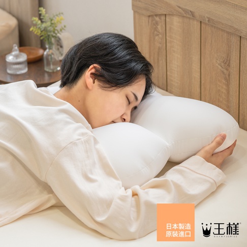 一位留著短髮的女性穿著淺色睡衣，側躺在床上使用王樣夢枕系列枕頭。枕頭呈現特殊的弧形設計以支撐頭部與手臂，女性雙手輕抱枕頭側邊，畫面採近景拍攝，呈現出舒適且放鬆的居家睡眠氛圍，右下角印有日文原裝進口說明與王樣品牌標誌。