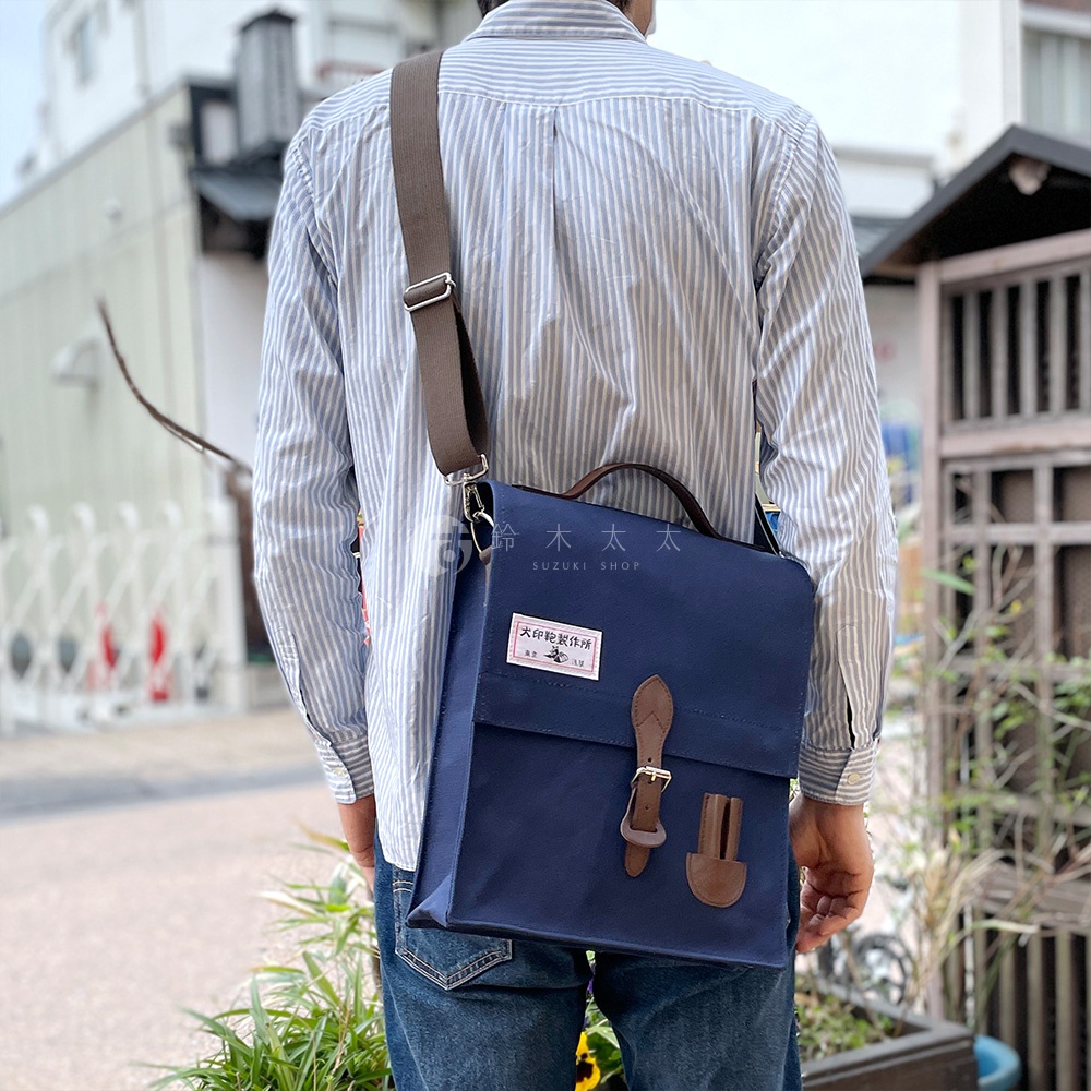 深藍色帆布直式側背包，配有棕色皮革提把與束帶扣環，包身有品牌日文標籤。