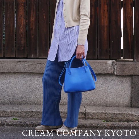 一位身穿藍色條紋襯衫、米色開襟毛衣和藍色寬褲的女士，手提一個亮藍色皮革托特包，包包有一條可拆卸肩帶。